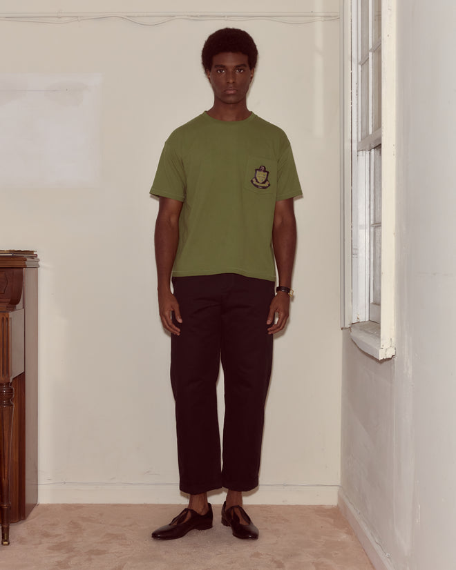A man in a green Theatre Patch Tee and black pants stands indoors by a window, facing the camera.