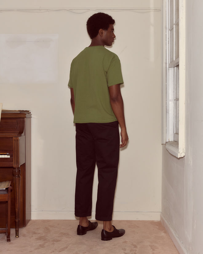 A man in a Theatre Patch Tee and black pants stands facing a corner near a piano and a window.