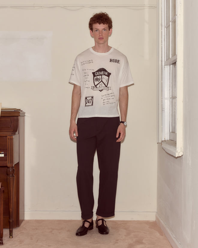 A person stands indoors near a window, wearing a Camp Crest Tee, black pants, and black shoes.