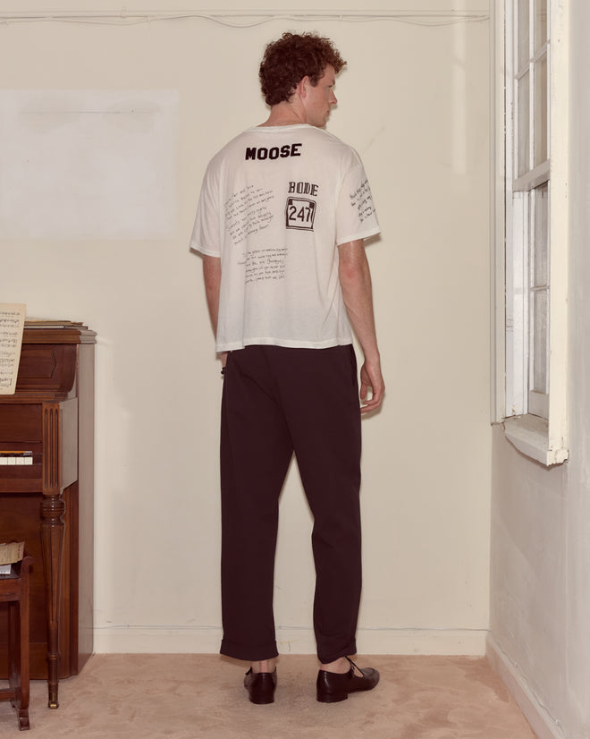 A man stands indoors facing away near a piano and window, wearing the Camp Crest Tee and black pants.