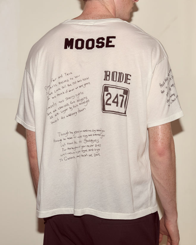 A person wearing the Camp Crest Tee, a 100% cotton white shirt featuring handwritten notes and "MOOSE" and "BODE 247" on the back.
