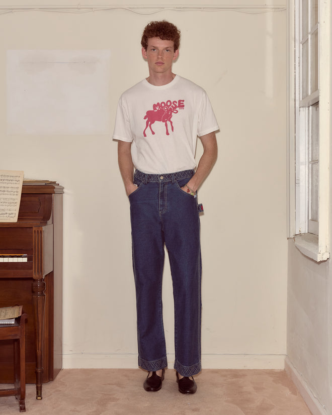 Indoors, a person in blue jeans stands by a piano, wearing the Antlers Tee—a pink shirt made in Portugal.