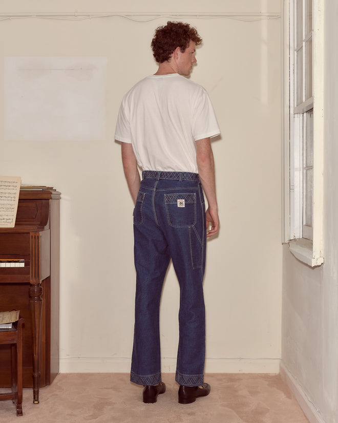 Man wearing the Antlers Tee, a midweight cotton pink shirt, with blue jeans stands facing a beige wall in a sparsely furnished room.