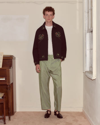 A man stands indoors by a piano, wearing the Lucky Draw Jacket, a hand-embroidered cotton piece, paired with a white shirt, green striped pants, and black shoes.