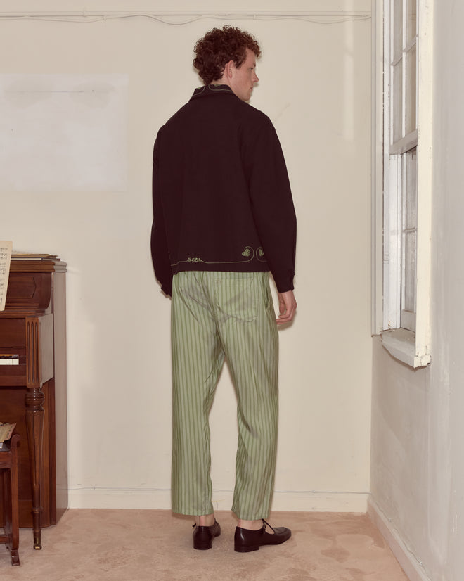 Indoors by a window and piano, a person faces away in green striped pants and the hand-embroidered Lucky Draw Jacket, showcasing a workwear-inspired silhouette.