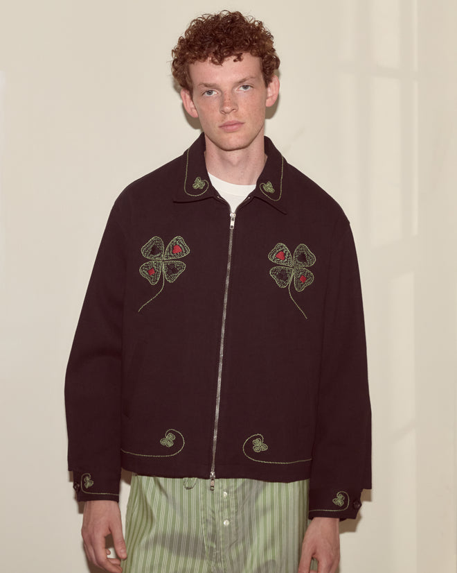 A young man stands against a plain wall, looking at the camera while wearing the Lucky Draw Jacket—a hand-embroidered cotton workwear piece featuring clover designs.