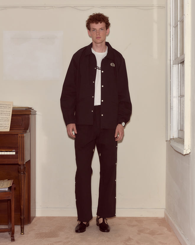 A man in a Twill Track Jacket - Black stands indoors by a piano and window, on a light carpeted floor.