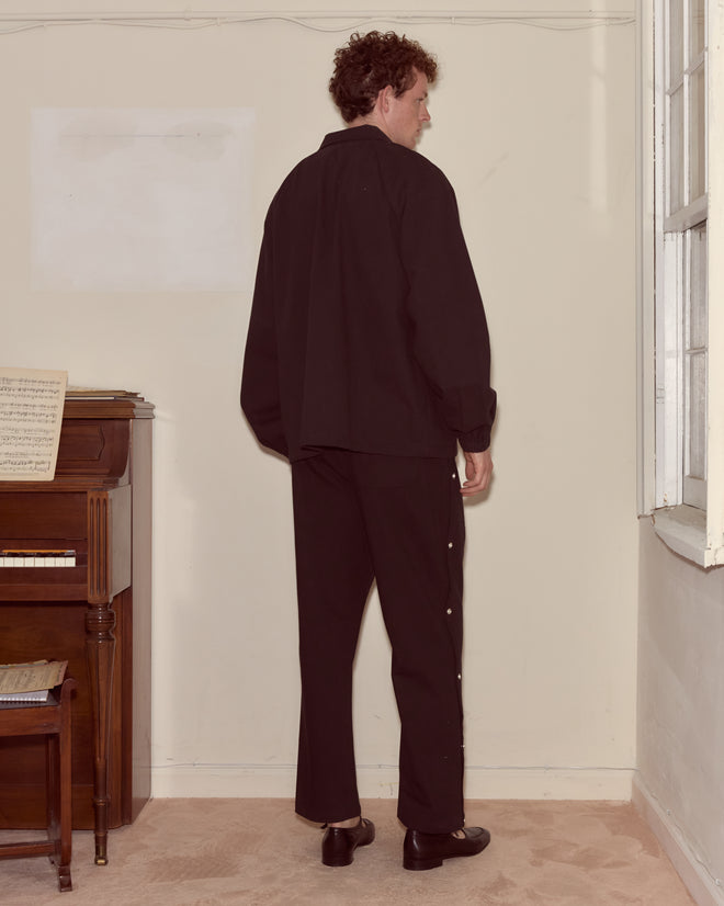 A person in a Twill Track Jacket - Black stands indoors near a piano, facing away from the camera.