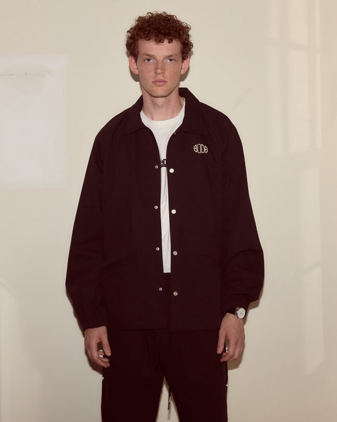 A young man with curly hair wears the Twill Track Jacket - Black over a white shirt, standing against a beige wall.