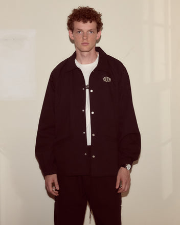 A young man with curly hair wears the Twill Track Jacket - Black over a white shirt, standing against a beige wall.