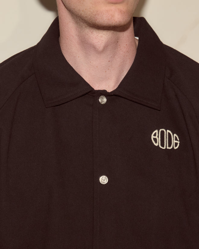 A person wearing the Twill Track Jacket - Black, a heavyweight cotton twill jacket with a black collar, Bode monogram, and "BODE" embroidered in white on the chest.