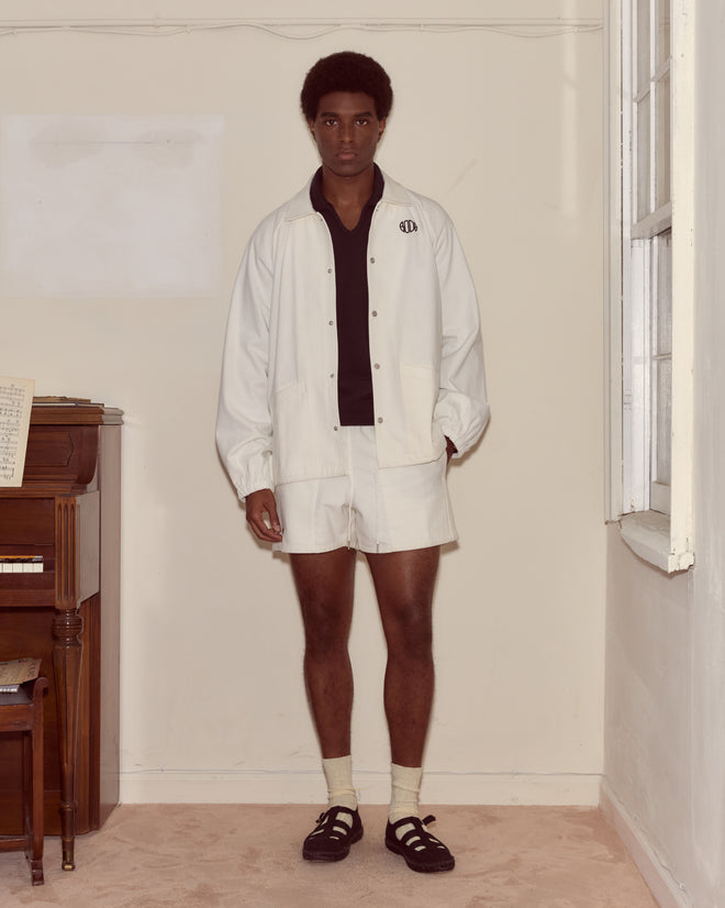 A man in a white Twill Track Jacket stands indoors by a piano and window, looking at the camera.