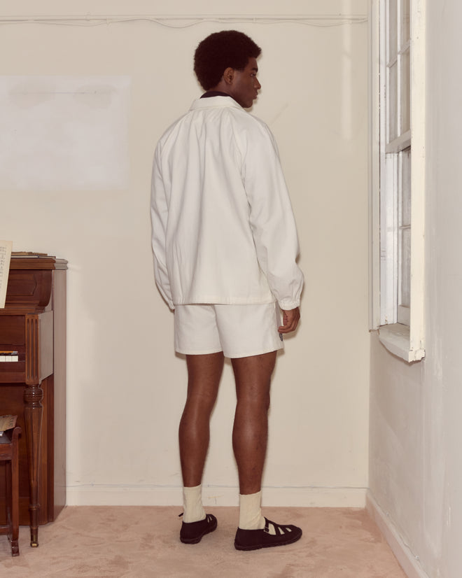 A person in a white shirt and shorts stands indoors by a window, facing away from the camera, with a Twill Track Jacket - White draped over one shoulder.