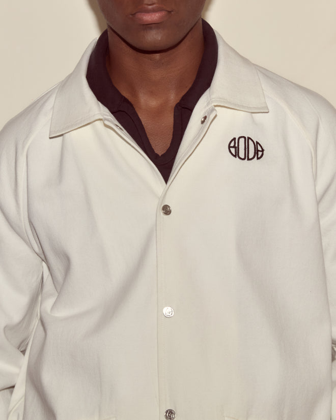 A person wearing the Twill Track Jacket - White, featuring a black logo, styled over a dark collared shirt.