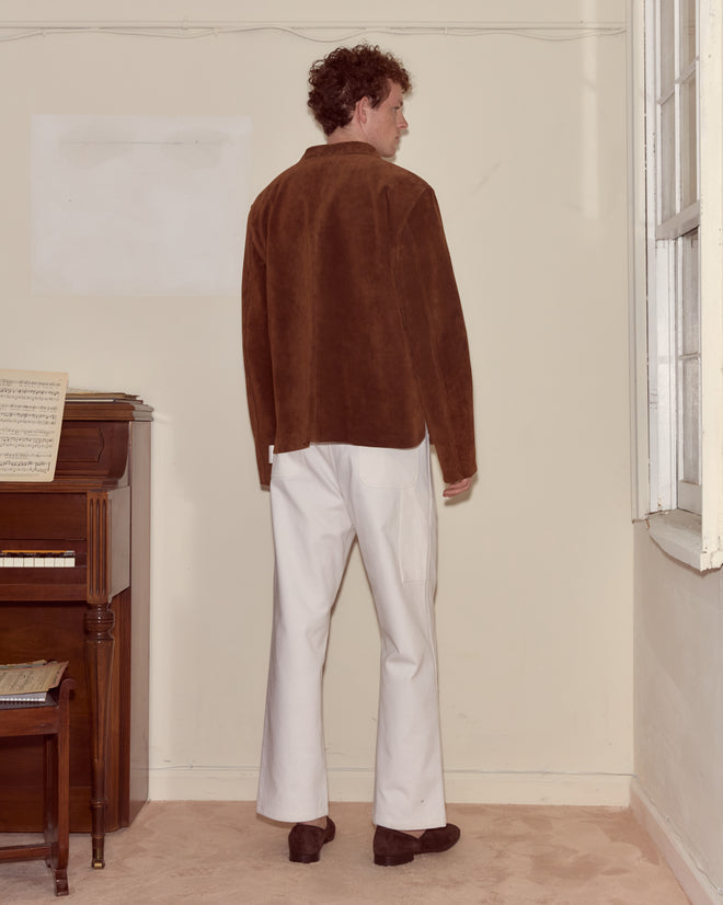 A man wearing the Suede Overshirt, featuring a heavyweight brown suede and workwear silhouette, pairs it with white pants while standing in a room with a piano and window, facing away.