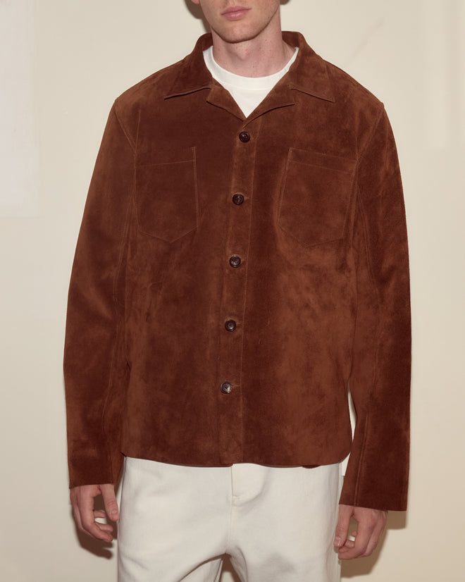 A person wears the Suede Overshirt, a heavyweight brown suede piece with a workwear silhouette, layered over a white shirt and white pants.