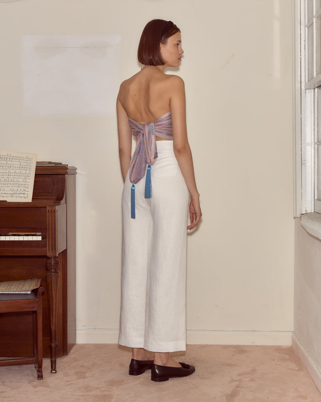 A woman in white pants and a backless top, draped in the blue and pink plaid Cheshire Scarf, made in Italy, stands by a piano near a window in a softly lit room.