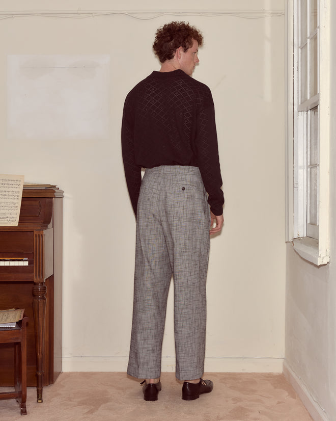 A man stands indoors facing a wall, wearing a black Anisette Polo and gray plaid trousers near a piano.