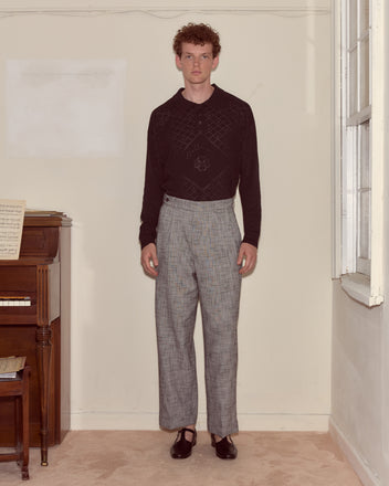 A man stands indoors by a piano, wearing the Anisette Polo, plaid pants, and black shoes.