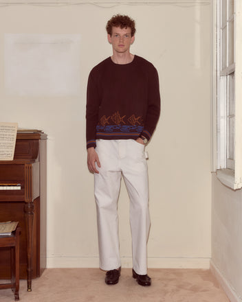 A man stands indoors wearing a Fish School Sweater, paired with white pants and black shoes.