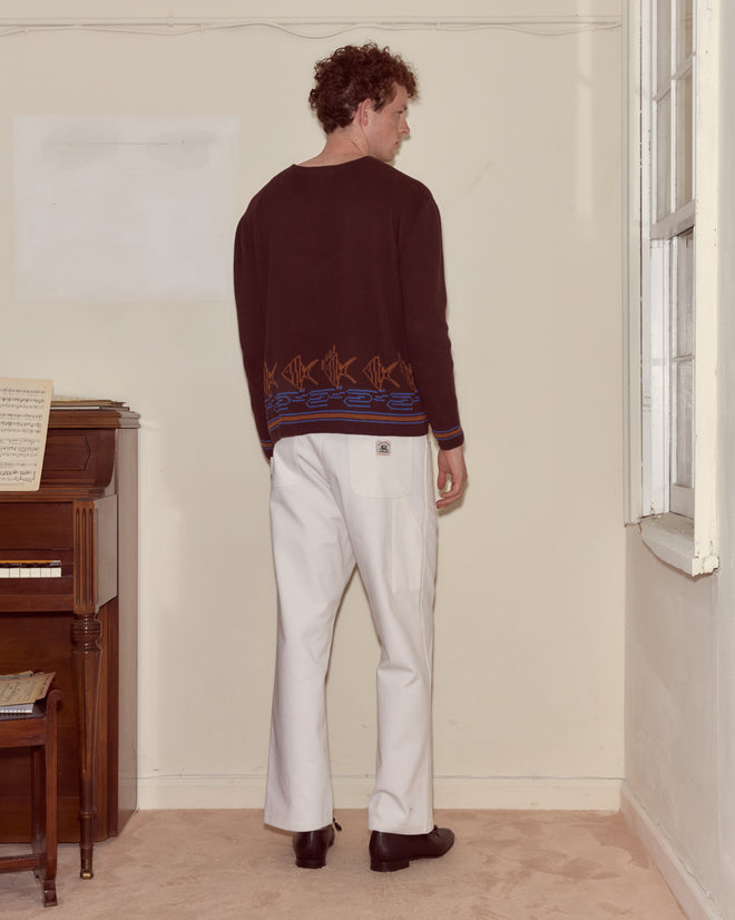 A man wearing the Fish School Sweater and white pants stands facing away in a room with a piano and sheet music.