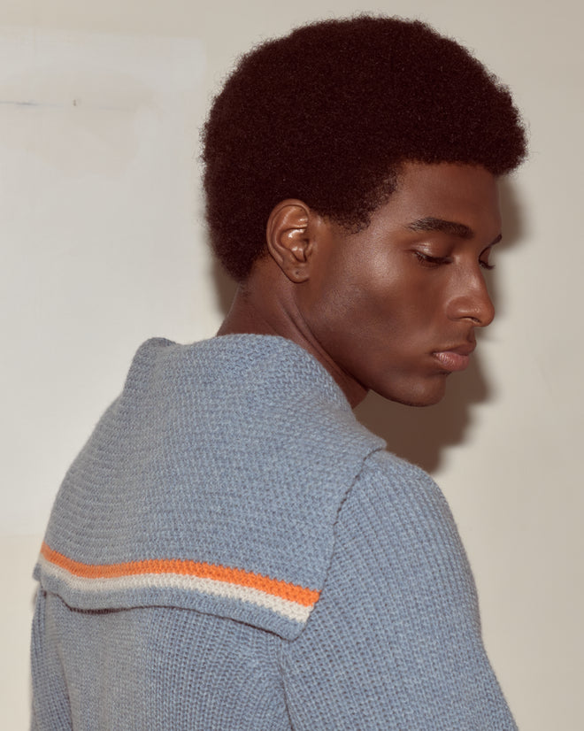 A person with short curly hair wears the Boat Deck Pullover, a blue virgin wool sweater with orange and white stripes and a sailor collar, looking downward.