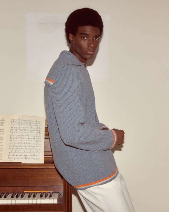 A man in a Boat Deck Pullover, featuring pirate ship buttons and blue virgin wool, stands by a piano with sheet music, glancing over his shoulder at the camera.