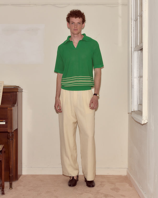 A man in a green Bentgrass Boxy Polo and cream trousers stands indoors beside a piano and a window.