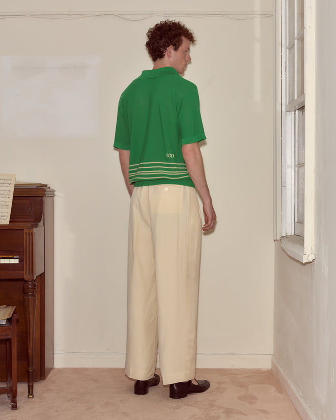 A man stands in a corner, facing away, wearing the green Bentgrass Boxy Polo with embroidered Bode and cream wide-leg trousers near a window and piano.