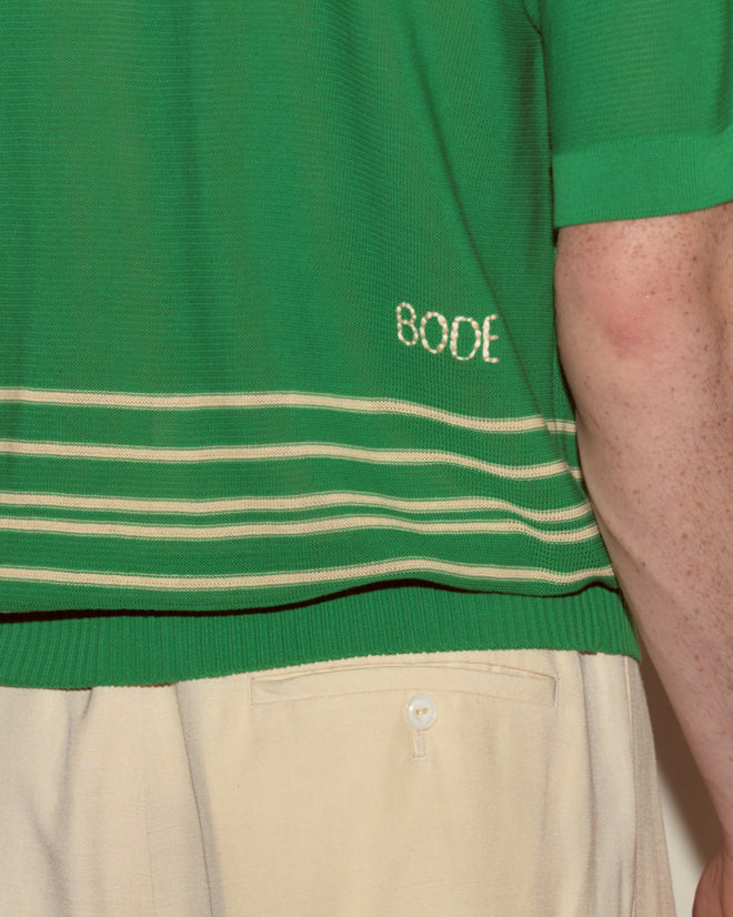 Person wearing a green Bentgrass Boxy Polo with an embroidered Bode logo and beige pants featuring a visible back pocket and button.