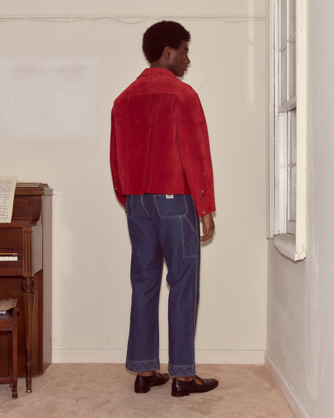 A man in a Welding Jacket - Red and blue jeans stands facing a window in a sparsely furnished room.