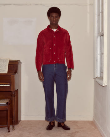 A man wearing a red Welding Jacket and blue jeans stands indoors by a piano and window, looking at the camera.