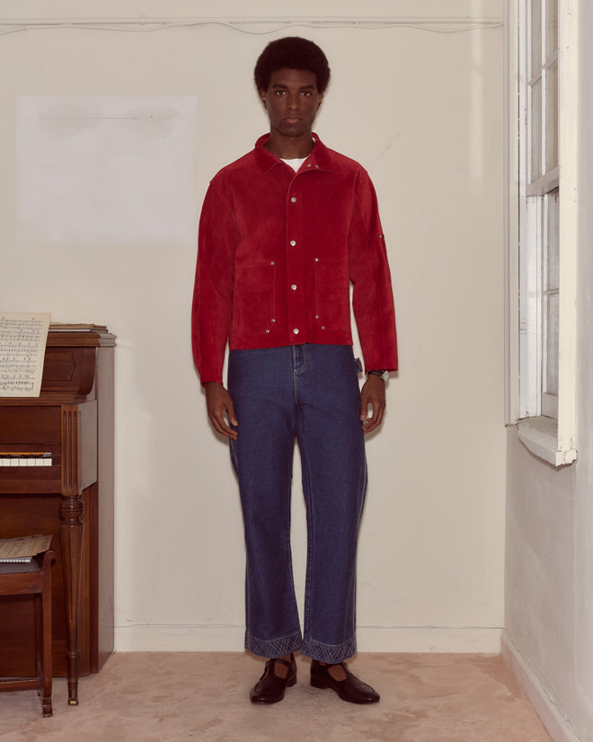 A man wearing a red Welding Jacket and blue jeans stands indoors by a piano and window, looking at the camera.