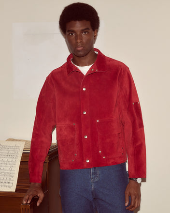 A man in a red Welding Jacket and jeans stands by a piano with sheet music on it, looking at the camera.