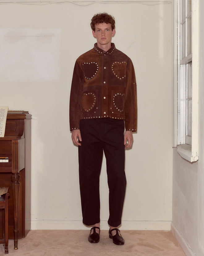 A man stands indoors wearing the Hearts Welding Jacket, a brown suede piece with studs and a heart appliqué, styled with black pants and black shoes.