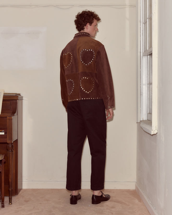 A person with curly hair stands indoors facing a wall, wearing the Hearts Welding Jacket and black pants.