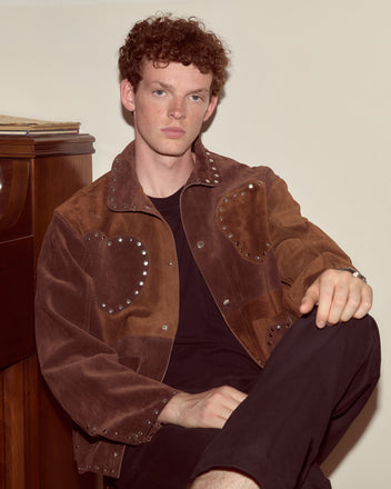 A man with curly red hair wears the Hearts Welding Jacket and sits by a wooden piano, looking at the camera.