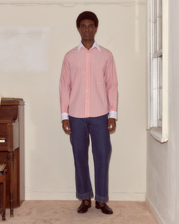 A person stands indoors in a pink striped Pickfair Shirt with a contrasting white collar, blue jeans, and black shoes next to a piano.