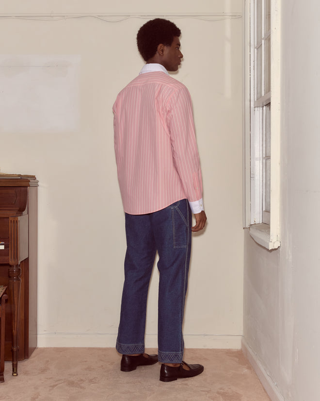 A person in a pink striped Pickfair Shirt with a contrasting white collar and jeans stands facing a corner window in a sparsely furnished room.