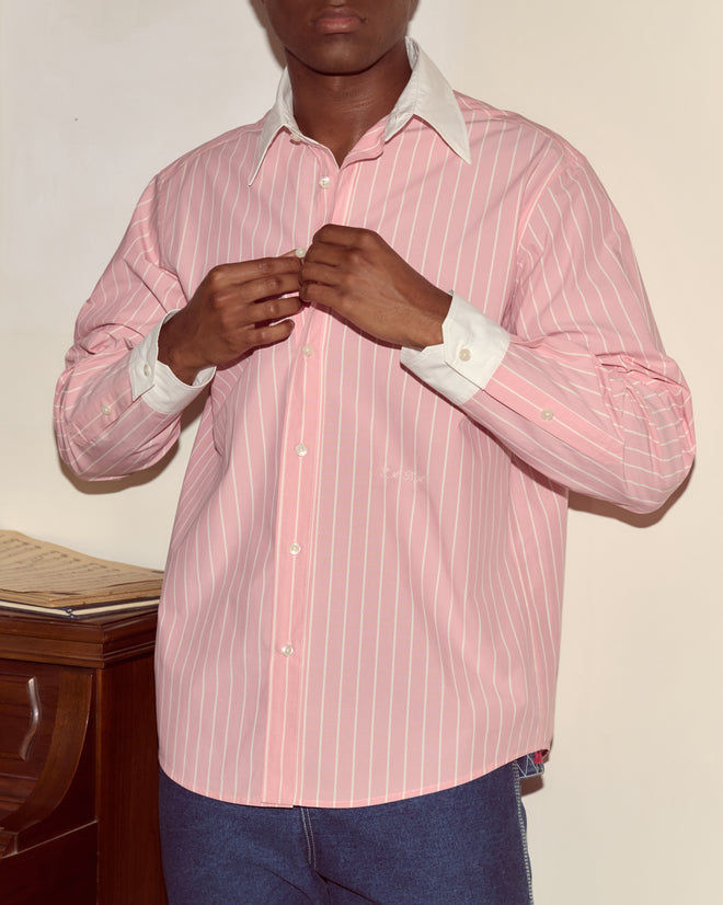 A person wearing a pink and white striped Pickfair Shirt with a contrasting white collar buttons it up while standing beside a wooden piano.