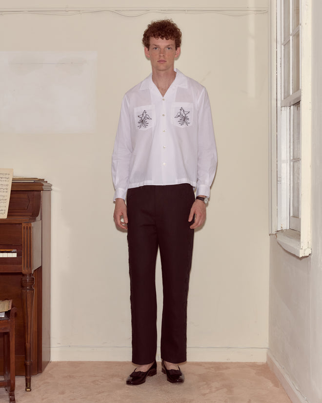 A person stands indoors by a piano wearing the Cockatoo Long Sleeve Shirt with black designs, paired with black pants and black heels.