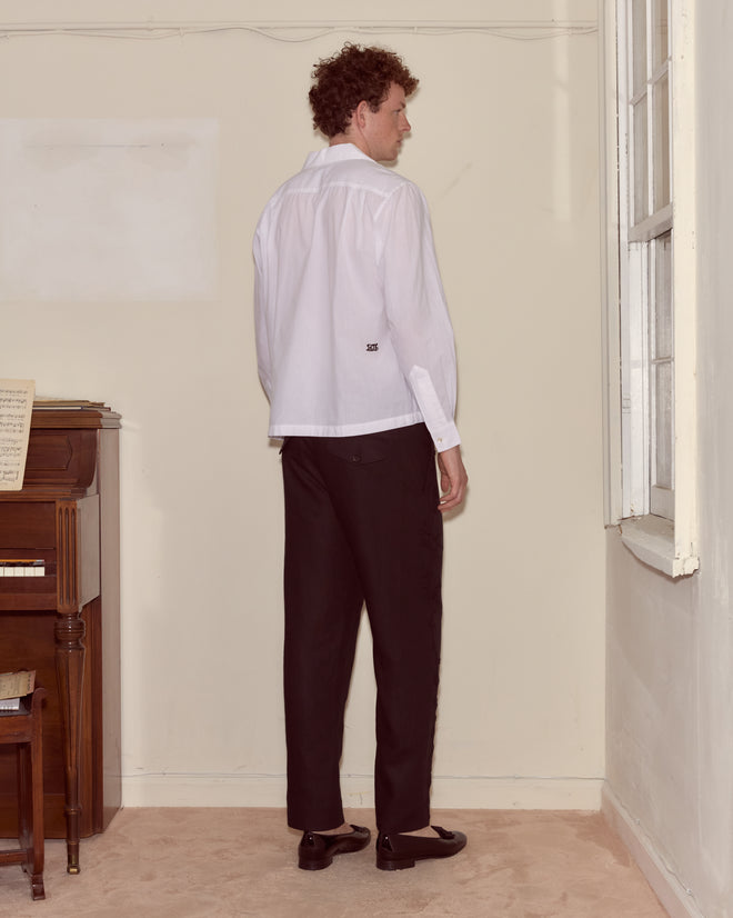 A man wearing the Cockatoo Long Sleeve Shirt and black pants stands facing a corner by a window, with a piano on his left.