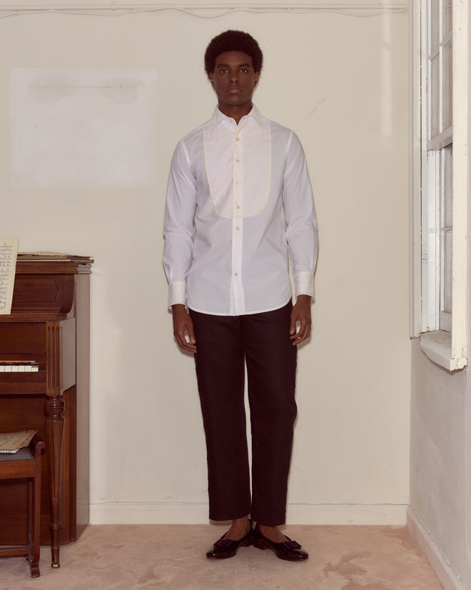 A man stands indoors by a piano, wearing the Closerie Tuxedo Shirt with ivory embroidery and black pants, looking straight at the camera.