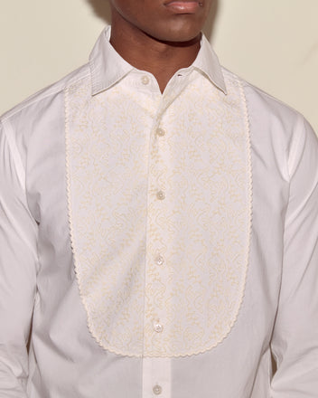 A person wearing the Closerie Tuxedo Shirt, a white cotton top featuring an embroidered, scalloped bib reminiscent of a classic tuxedo shirt.