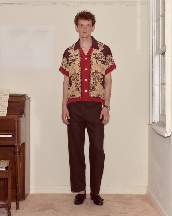 A man in a Storybook Short Sleeve Shirt and dark pants stands by a piano in a sunlit room.
