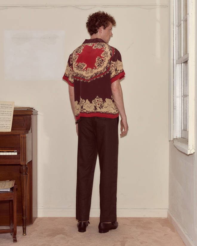 A man in a Storybook Short Sleeve Shirt and dark pants stands facing a wall near a piano in a softly lit room.