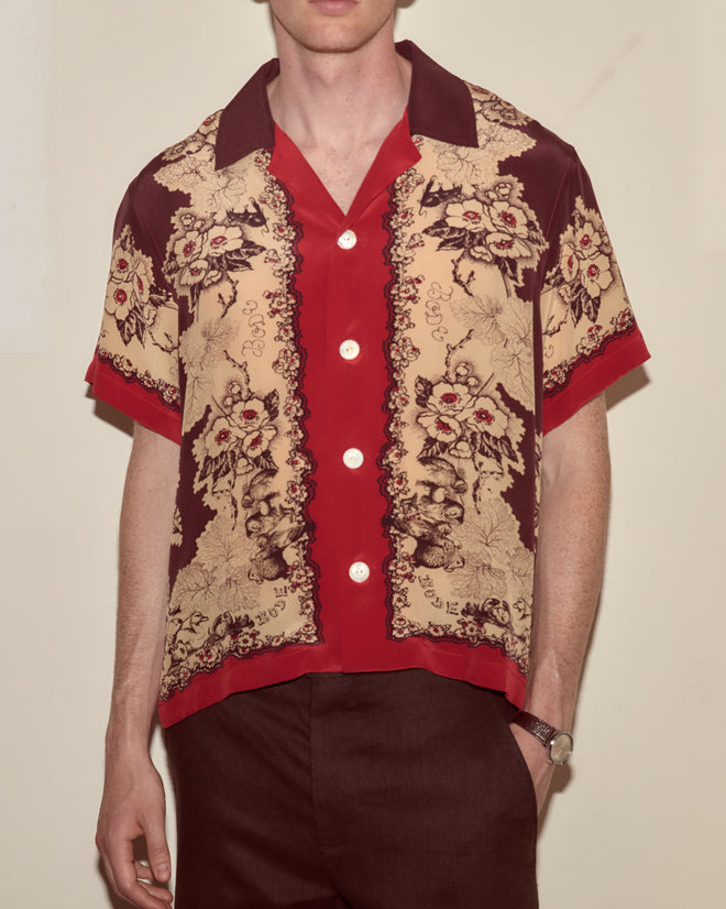 A man wears the Storybook Short Sleeve Shirt, featuring vintage-inspired red and beige Victorian floral patterns, paired with brown pants.