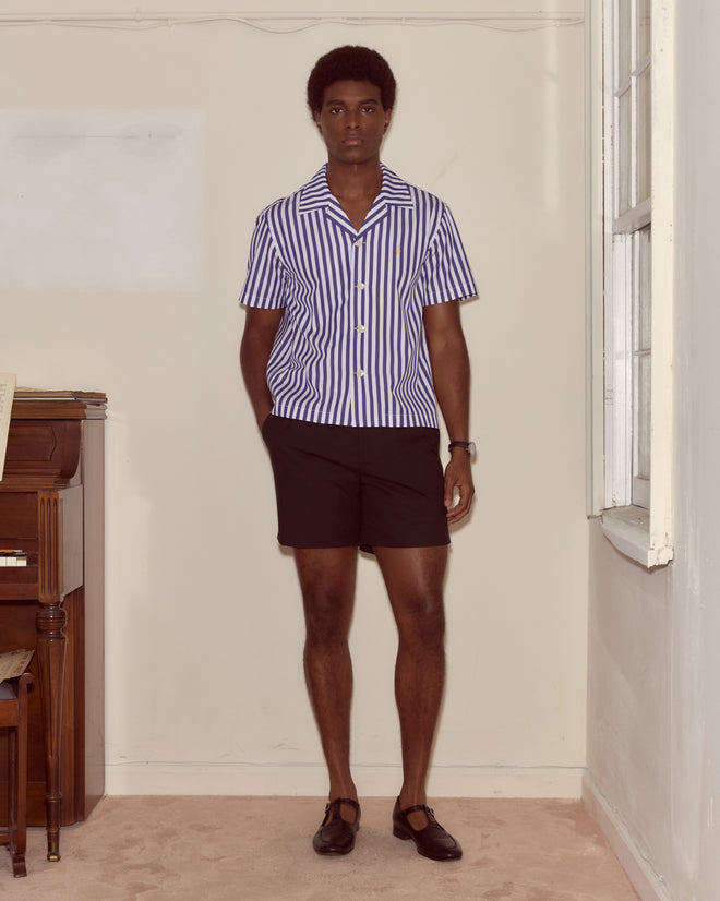 Indoors near a piano and window, a person wears the Calliope Short Sleeve Shirt featuring 1930s-inspired indigo and white stripes, paired with black shorts and black shoes.