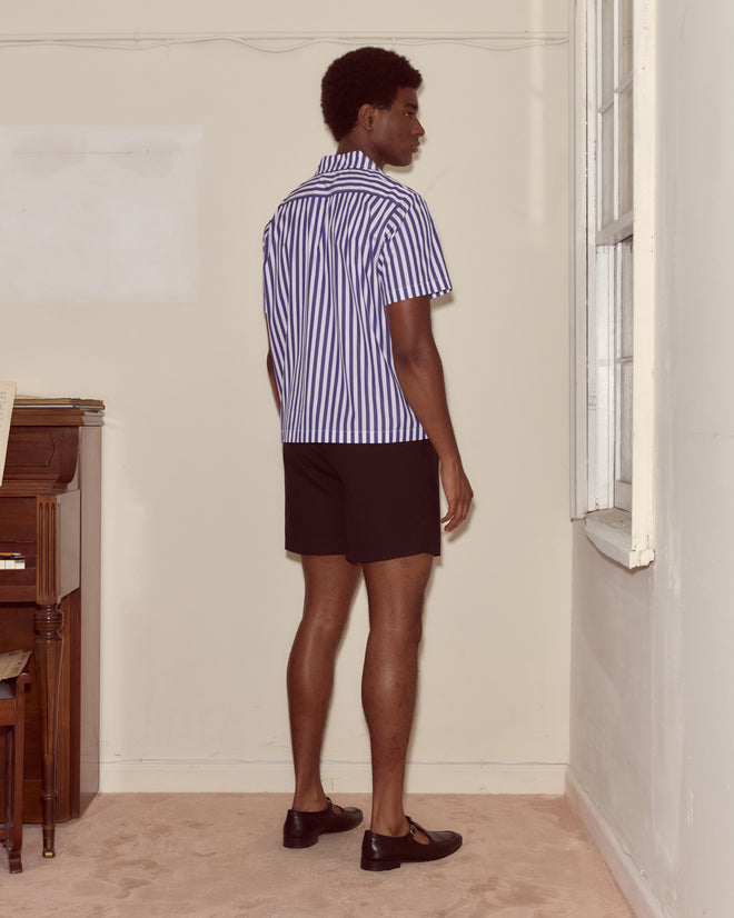 A man wearing the Calliope Short Sleeve Shirt, featuring indigo and white stripes, and black shorts stands by a window in a beige room with a piano.