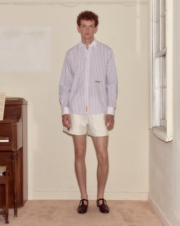 A man in a lightweight Steamboat Stripe Shirt with a contrasting white collar and white shorts stands indoors by a piano, wearing black shoes.